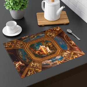 May include: Rectangular placemat featuring a Renaissance-style ceiling painting with figures in shades of gold, blue, and brown. The placemat has a dark border and the text "Savor Smith" in the lower right corner. A white teacup and saucer are on the left.