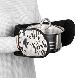 May include: A black and white oven mitt with a dalmatian print design holds a stainless steel saucepan with a glass lid. The mitt has a quilted texture and is designed to protect hands from heat.