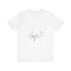 Könnte beinhalten: Weißes T-Shirt mit dem Wort "love" in handschriftlicher Schrift, begleitet von einem kleinen Herzsymbol. Das Design ist in Dunkelgrau gehalten. Das Shirt hat kurze Ärmel und einen Rundhalsausschnitt.