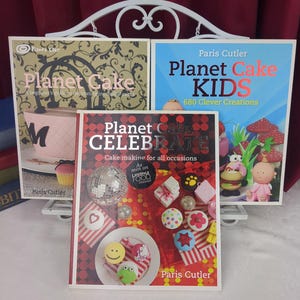 以下が含まれることがあります： パリス・カトラー著の「Planet Cake」、「Planet Cake KIDS 680 Clever Creations」、「Planet Cake CELEBRATE」というタイトルの3冊の本。表紙には、白いディスプレイスタンドの上に飾られた、デコレーションケーキ、カップケーキ、フォンダンフィギュアのカラフルな写真が掲載されています。