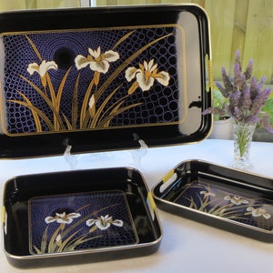 May include: A set of three rectangular black serving trays with gold-toned handles and edges. Each tray features a central graphic of white and gold iris flowers set against a dark purple background with a repeating pattern of concentric circles.