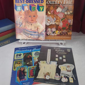 Puede incluir: Cuatro revistas vintage de manualidades y tejido exhibidas en un soporte de alambre blanco. Los títulos incluyen Family Circle Best-Dressed Baby, Country Fair Food & Crafts, The Australian Wool Corporation Knitting Book y Peter Rabbit Counts con varios proyectos.