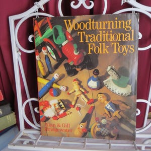 May include: A book titled "Woodturning Traditional Folk Toys" with a colorful cover featuring various wooden toys. The book is displayed on a white metal shelf, with other books visible on the left. The cover includes the authors' names.