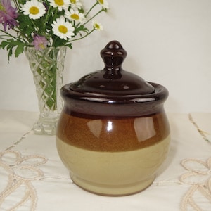 May include: A brown and beige ceramic sugar bowl with a lid. The lid has a knob on top. The bowl is sitting on a white tablecloth with lace trim.