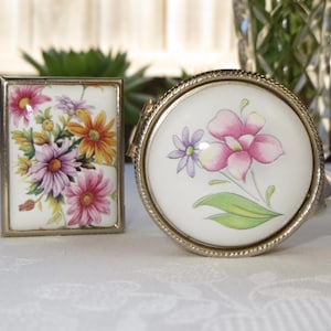May include: Two vintage floral trinket boxes with silver metal frames. The box on the left has a white background with pink, yellow, and purple flowers. The box on the right has a white background with a pink flower and green leaves.