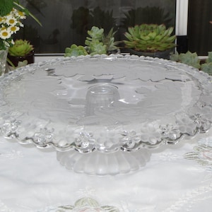 May include: A clear glass cake stand with a frosted base and a scalloped edge. The top of the stand features a delicate floral design.