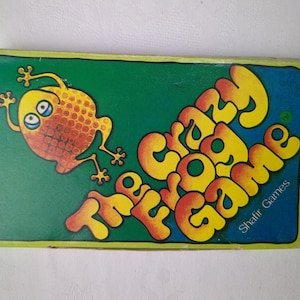 May include: A rectangular board game box with a green background featuring a yellow and orange cartoon frog illustration. The text reads The Crazy Frog Game and Shafir Games in large, stylised yellow and orange letters across the front cover.