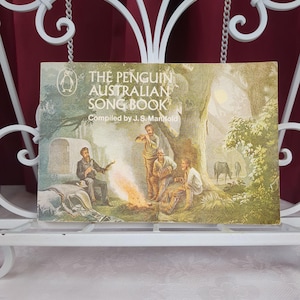 May include: The Penguin Australian Song Book compiled by J. S. Manifold is a softcover book featuring an illustration of four people sitting around a campfire in a wooded area with a wagon and horses in the background, all in muted green tones.