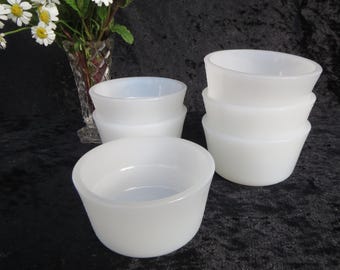 May include: A collection of white, opaque glass bowls. The bowls are stacked in groups of two and three, and are also shown individually. A vase of flowers is in the background.