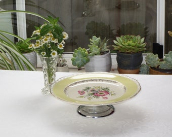 Op de afbeelding: Een vintage taartplateau met een gele rand en een bloemenmotief. Het plateau heeft een zilveren voet en staat op een wit tafelkleed.