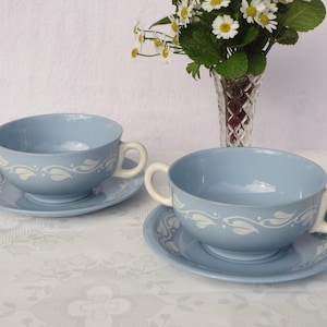 May include: Two light blue ceramic soup bowls with matching saucers. Each bowl has two handles and a white floral design. A vase of white flowers sits in the background.
