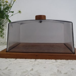 May include: A wooden serving tray with a clear plastic dome lid. The lid has a wooden knob on top. The tray is on a white crocheted surface.