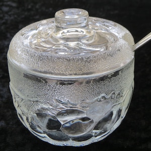 Peut inclure: Un bol à sucre en verre transparent avec un couvercle et une cuillère. Le bol a un motif texturé de feuilles et de fruits.