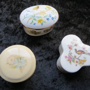 May include: Three porcelain trinket boxes with floral and bird designs. The boxes are white with gold trim and feature delicate floral patterns and a bird on one of the boxes.