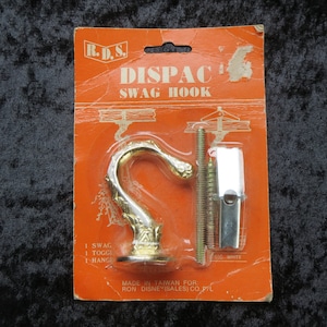 Könnte beinhalten: Ein goldfarbener Swag-Haken mit dekorativem Design. Der Haken ist auf einer leuchtend orangefarbenen Karte verpackt mit dem Text "DISPAC SWAG HOOK" und "MADE IN TAIWAN FOR RON DISNEY(SALES) CO. P/L".