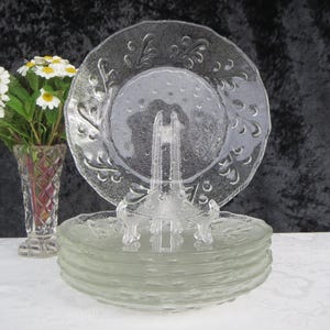 May include: A stack of clear glass plates with a textured, embossed design. The plates are displayed on a clear plate stand. A clear glass plate with a similar design is on the stand. A vase of white flowers is to the left.