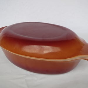 Puede incluir: Una fuente ovalada marrón con tapa a juego. La fuente tiene un acabado brillante y asas integradas a cada lado. El color pasa de un naranja más claro en la base a un marrón más oscuro en la parte superior.