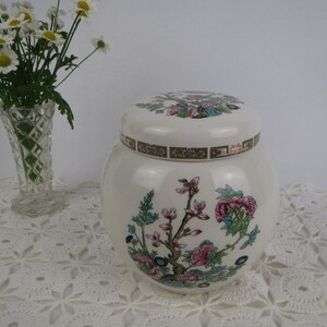 May include: White ceramic jar with a lid, featuring a floral design with pink and green flowers. The jar has a gold trim around the top.