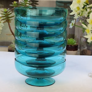 May include: A stack of six teal glass dessert bowls. Each bowl has a rounded base and a stemmed foot. The bowls are arranged on a white surface, with flowers and plants in the background.