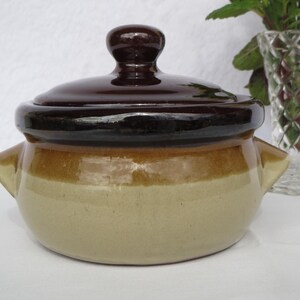 Puede incluir: Una cazuela de cerámica con tapa. El plato es de color beige claro con una banda marrón alrededor del centro y una tapa y asa de color marrón oscuro. Tiene dos asas pequeñas a cada lado.