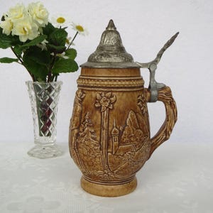 Puede incluir: Una jarra de cerveza de cerámica marrón con un diseño en relieve detallado de un paisaje montañoso, árboles y un edificio. Tiene una tapa y un asa articuladas de metal. Un jarrón de cristal transparente con flores está al lado.