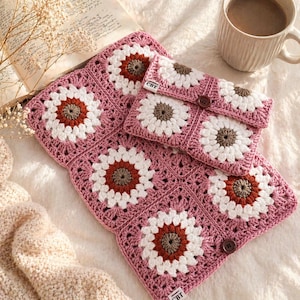 Pode incluir: Um conjunto de crochê feito à mão com um padrão de margarida, apresentando um cobertor rosa e uma clutch combinando. O design floral incorpora detalhes em branco, marrom e laranja. A clutch tem um fecho de botão e o cobertor tem as letras "CBT".