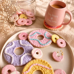 May include: A selection of colourful crochet donuts, including pink, yellow, and purple, displayed on a plate. A pink mug with the words "Stay Happy Stay Cosy" is on a crocheted coaster. A glass jar contains mini donuts. Measurements are in cm.
