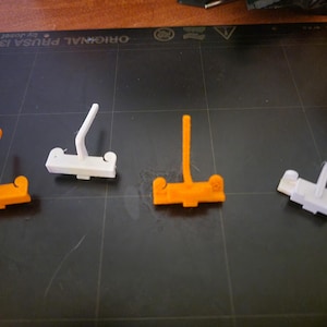 Puede incluir: Cuatro objetos de plástico impresos en 3D. Dos son naranjas y dos son blancos. Cada uno tiene una base con un poste vertical y un brazo curvo. Los objetos están dispuestos sobre una superficie oscura.