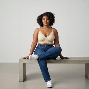 Könnte beinhalten: Ein beiges Spitzen-Bralette mit V-Ausschnitt und gewellten Kanten, kombiniert mit einer dunkelblauen Jeans und weißen Sneakern. Das Bralette zeichnet sich durch ein zartes Blumenmuster, dünne Träger und eine bauchfreie Passform aus.