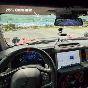 Puede incluir: Vista interior de un Ford Bronco rojo con un volante negro, un tablero digital y un espejo retrovisor con una cámara de salpicadero. El parabrisas tiene un tinte cerámico del 25%.