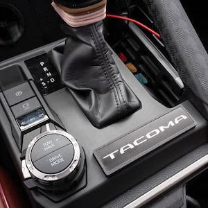 May include: Close-up of a car's interior console. Features a black gear shift, buttons for parking brake, 4L/4H/2H, and a circular dial with "TOW HAUL" and "DRIVE MODE" options. The console also has a "TACOMA" emblem.