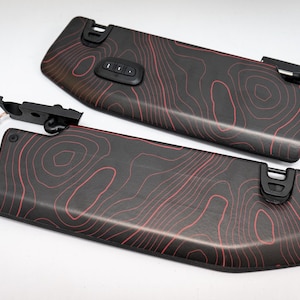 May include: Two black car sun visors with a red topographic pattern. One visor has a control panel with buttons. The visors have black mounting hardware and a wire with connectors.