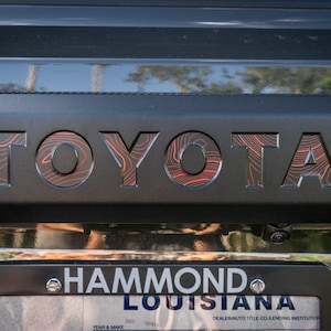 May include: A black Toyota truck with a number plate that reads "HAMMOND LOUISIANA". The Toyota logo is a topographic map design with red lines.