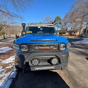 Peut inclure: Un Toyota FJ Cruiser bleu avec un pare-chocs avant noir et une calandre noire. Le véhicule est équipé d'un porte-bagages de toit avec une barre lumineuse et deux phares LED ronds sur le pare-chocs avant. Le logo Toyota est visible sur la calandre.