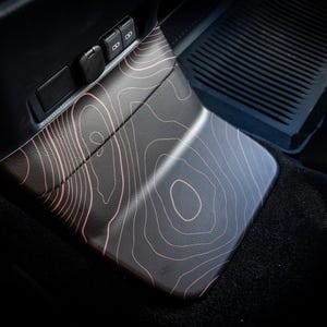 以下が含まれることがあります： 黒い車のインテリアで、センターコンソールに地形図のパターンが施されています。コンソールは木目調で、茶色と黒の配色になっています。