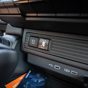 以下が含まれることがあります： 車のインテリアのクローズアップで、2つのボタンが付いた黒いパネルが表示されています。左側のボタンには「VIEW」、右側のボタンには「SAFETY SEAT-O」と表示されています。また、USBポートが2つと充電ポートも付いています。