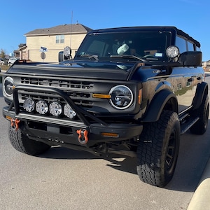 Puede incluir: Un Ford Bronco negro con un parachoques delantero negro y una jaula antivuelco negra. El vehículo tiene una parrilla negra con un logotipo Ford amarillo y un capó negro. El vehículo tiene un techo negro y espejos laterales negros. El vehículo tiene ruedas negras con neumáticos negros. El vehículo tiene un parachoques delantero negro con un cabrestante negro y un gancho de remolque negro. El vehículo tiene un parachoques delantero negro con una barra de luces negra.