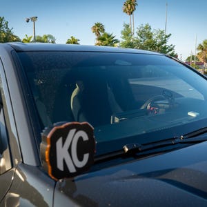 Puede incluir: Primer plano de un SUV gris con capó y parabrisas negros. Una luz KC negra y naranja está montada en la parte delantera. El interior del coche es visible a través del parabrisas, con el volante y el salpicadero a la vista.