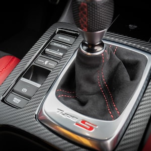 May include: Close-up of a black leather gear shifter with red stitching and a carbon fibre trim panel. The panel has buttons for "Brake Hold", "Eco", "Sport", and "Individual" driving modes. The gear shifter has a "Type S" logo.