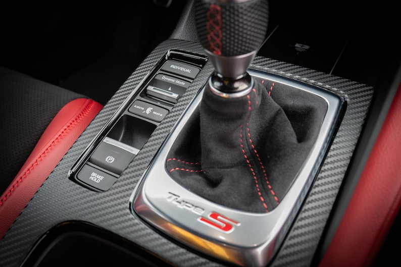 May include: Close-up of a black leather gear shifter with red stitching and a carbon fibre trim panel. The panel has buttons for "Brake Hold", "Eco", "Sport", and "Individual" driving modes. The gear shifter has a "Type S" logo.