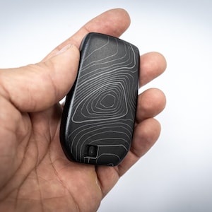 Op de afbeelding: Een zwarte sleutelhanger met een topografisch kaartontwerp in witte lijnen. De sleutelhanger wordt in een hand gehouden tegen een witte achtergrond. De sleutelhanger heeft een gladde, matte afwerking.