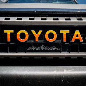 Puede incluir: Primer plano de una parrilla de coche negra con la palabra "TOYOTA" en letras amarillas y naranjas. La parrilla tiene un gráfico de cordillera y está rodeada por un marco plateado. El parachoques del coche es negro.