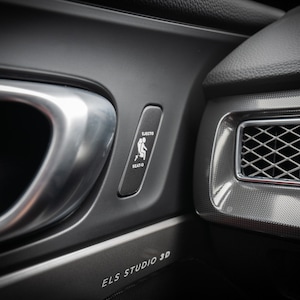 Puede incluir: Primer plano del interior de un coche con una manija de la puerta plateada, un botón negro con un icono blanco de una persona sentada en un asiento y el texto "ELS STUDIO 3D".