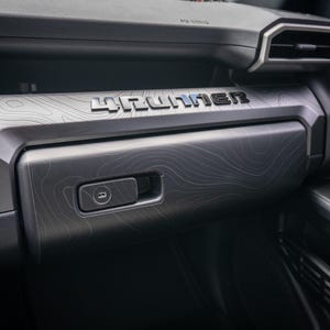 以下が含まれることがあります： 車両のダッシュボードのインテリア詳細。ダッシュボードは、グレーと黒の色合いのテクスチャ加工された地形パターンが特徴です。「4RUNNER」の文字が銀色で表示されています。ハンドル付きのグローブボックスが見えます。