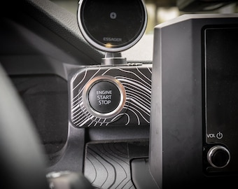 Peut inclure: Gros plan sur l'intérieur d'une voiture noire avec un bouton de démarrage/arrêt du moteur argenté. Le bouton présente un motif topographique blanc et noir et les mots "Engine Start Stop" sont imprimés dessus.