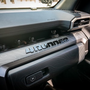 May include: Interior view of a vehicle's dashboard. The dashboard is primarily dark gray with a topographic pattern. The text "4Runner" is prominently displayed in silver lettering. Air vents and other controls are visible.
