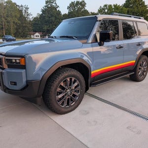 Puede incluir: Un SUV azul claro con techo blanco y detalles negros. El vehículo tiene una franja retro en rojo, amarillo y naranja en el lateral. Está estacionado sobre una superficie de hormigón.