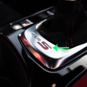 May include: Close-up of a black leather car gear shifter with red stitching. The gear shifter has a silver trim with the word "Sport" and the letter "S" in red.