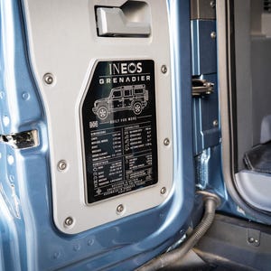 May include: A light blue vehicle door interior featuring a black metal plaque with the Ineos Grenadier logo, a line drawing of the vehicle, and technical specifications including dimensions, powertrain details, and off-road capability metrics.
