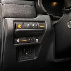 May include: Close-up of a car's dashboard with various buttons and controls. The buttons are labeled "KC", "OFF", "AUDIO", and have icons for a car, a person, and a smiley face.